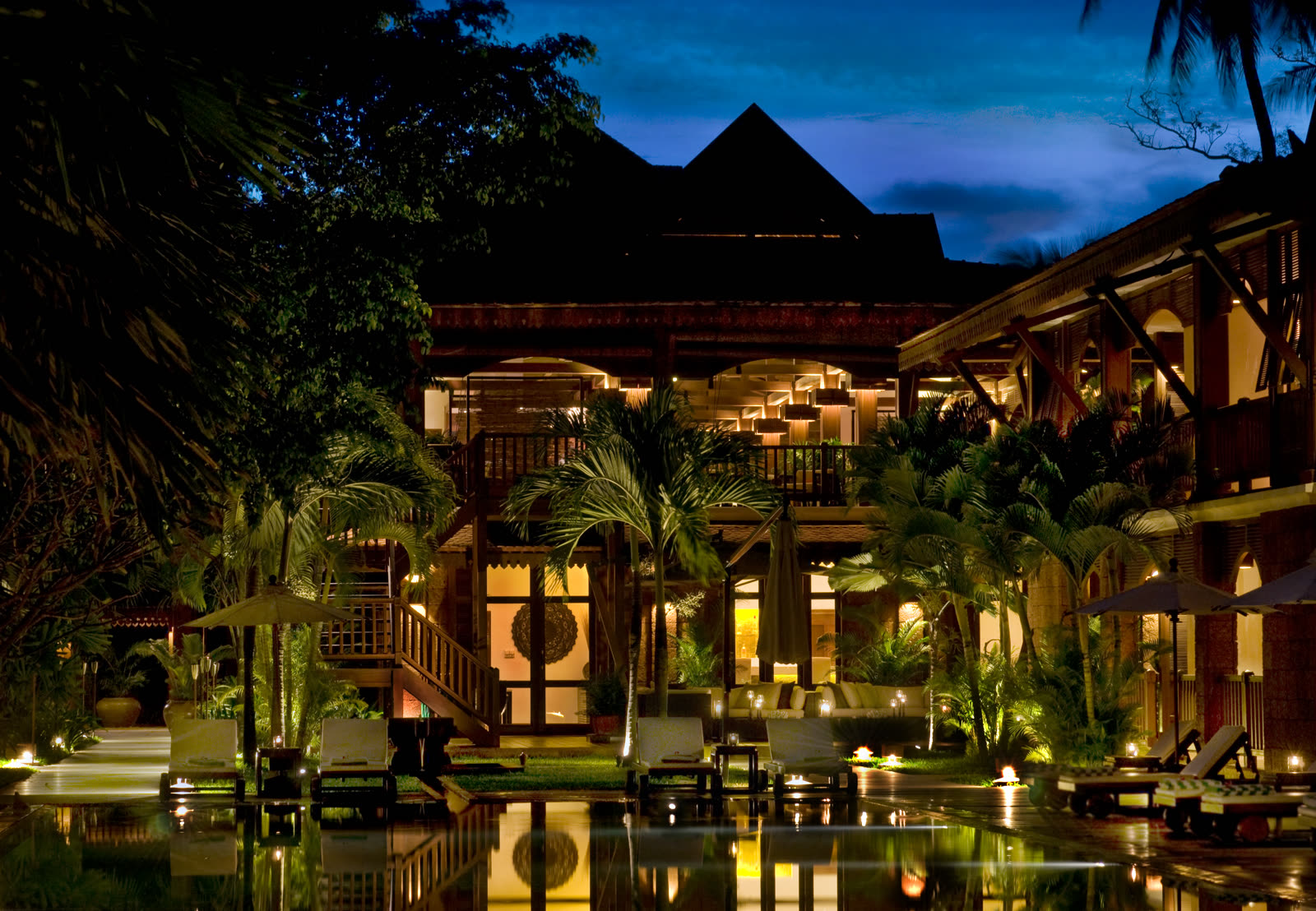 Timber pavilions lit at dusk beside a pool among palms